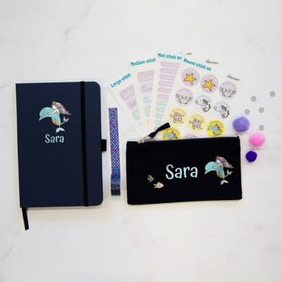 Mermaid back to school mega bundle. Including four sheets of stick on name labels, a blue notebook with a picture of a mermaid riding a dolphin and the name in light blue, a black pencil case with name in light blue, picture of mermaid riding dolphin to right of text and two fish on the left of text.