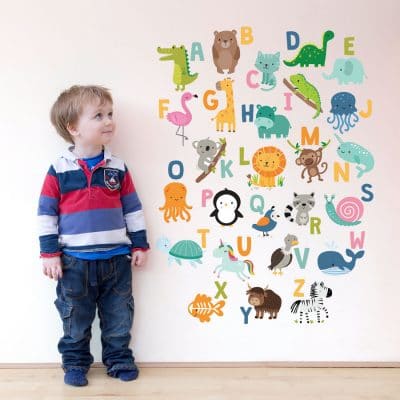 Jungle Alphabet Wall Sticker, jungle wall stickers. Image shows a colourful alphabet sticker arranged in a rectangular format with lots of animals. The sticker has been displayed on a plain wall next to a small child.
