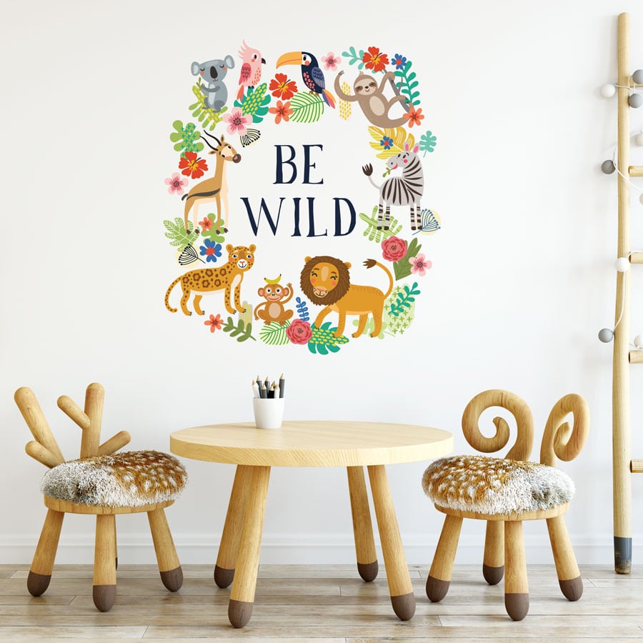 Be wild wall sticker, jungle wall stickers. Image shows The text saying "Be Wild" in large black letters surrounded by a frame of animals and plants. The animals included are, a lion, a giraffe, a cheetah, a koala, a monkey, a sloth, a gazelle and two birds. Sticker is displayed above a wooden table with wood chairs carved to look like antlers.