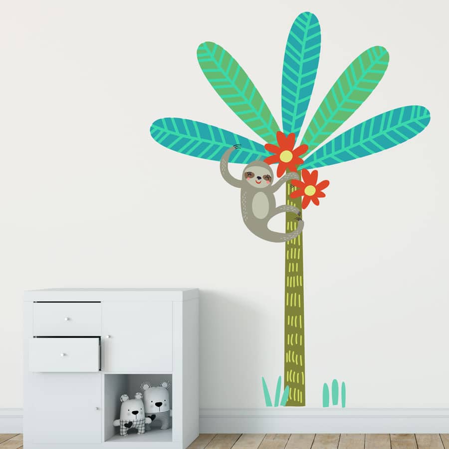 Sloth and tree wall sticker. Image shows a large slender tree with big green leaves and red flowers as well as a sloth hanging from the upper part of the tree. At the bottom of the tree there are some light green grass shaped stickers. This wall sticker has been placed on a plain wall next to a chest of drawers.