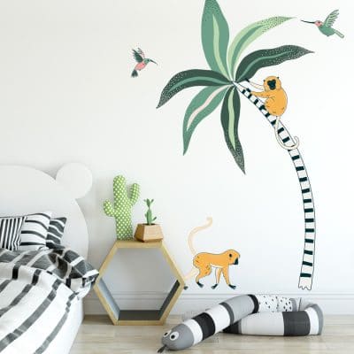 Tropical Palm And Monkeys Wall Stickers. Image displays a room with a large stripy palm tree sticker with green leaves, a yellow monkey on the tree and one walking along the ground, and two pink and green humming birds on the wall.