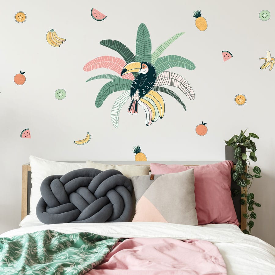 Tropical toucan wall sticker pack, jungle wall sticker. Sticker is of a toucan sat in the centre of green and pink palm leaves. Surrounding the main sticker is colourful fruit, pineapples/bananas/melon/oranges. The sticker has been placed in a bedroom above a green and pink bedspread.