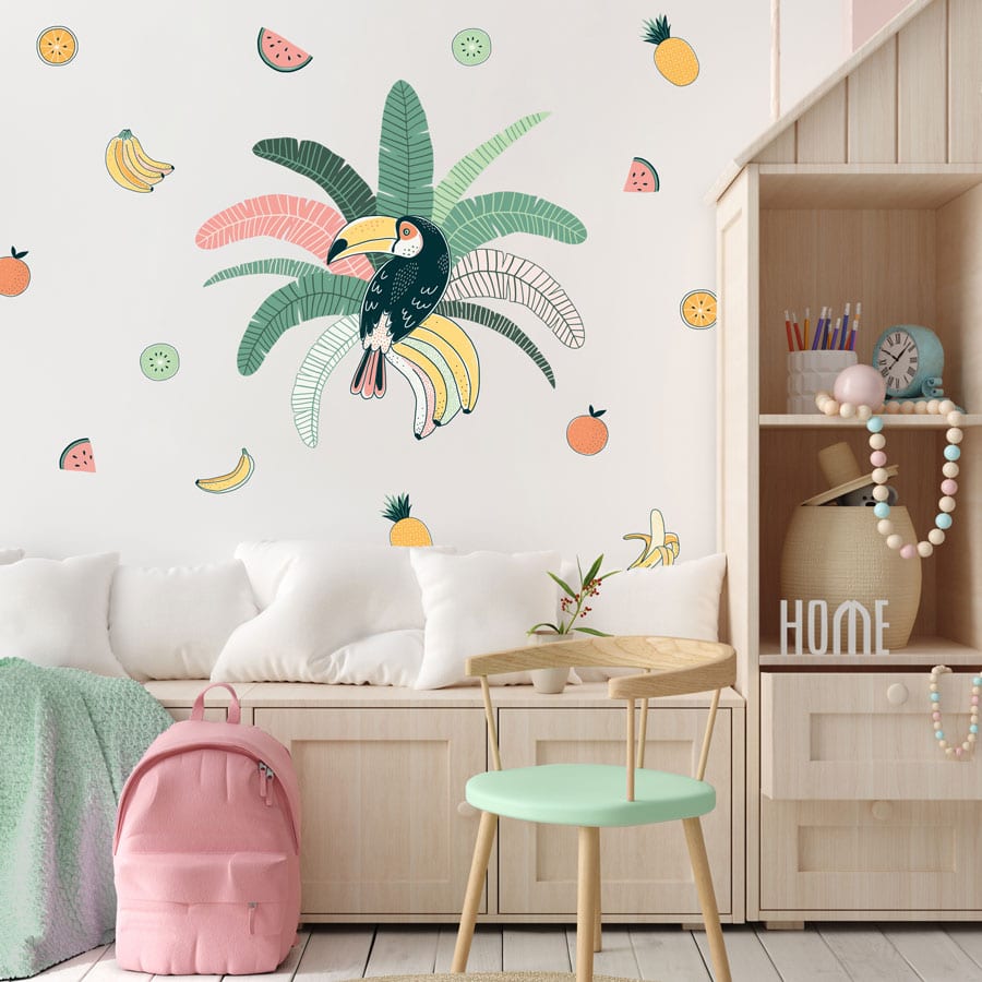 Tropical toucan wall sticker pack, jungle wall sticker. Sticker is of a toucan sat in the centre of green and pink palm leaves. Surrounding the main sticker is colourful fruit, pineapples/bananas/melon/oranges. Sticker has been placed in a bedroom above a low wooden unit and next to tall shelves.