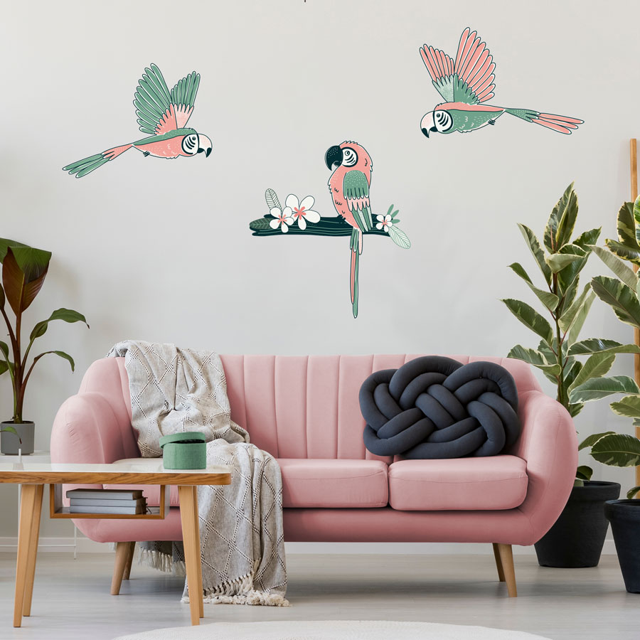 Tropical parrot wall sticker pack, jungle wall stickers. Image shows three pink and mint green parrot stickers, two are flying and one is sat on a dark green branch with a white flower. The stickers have been placed on a white wall above a pinka sofa with green, grey and natural toned furnishings.