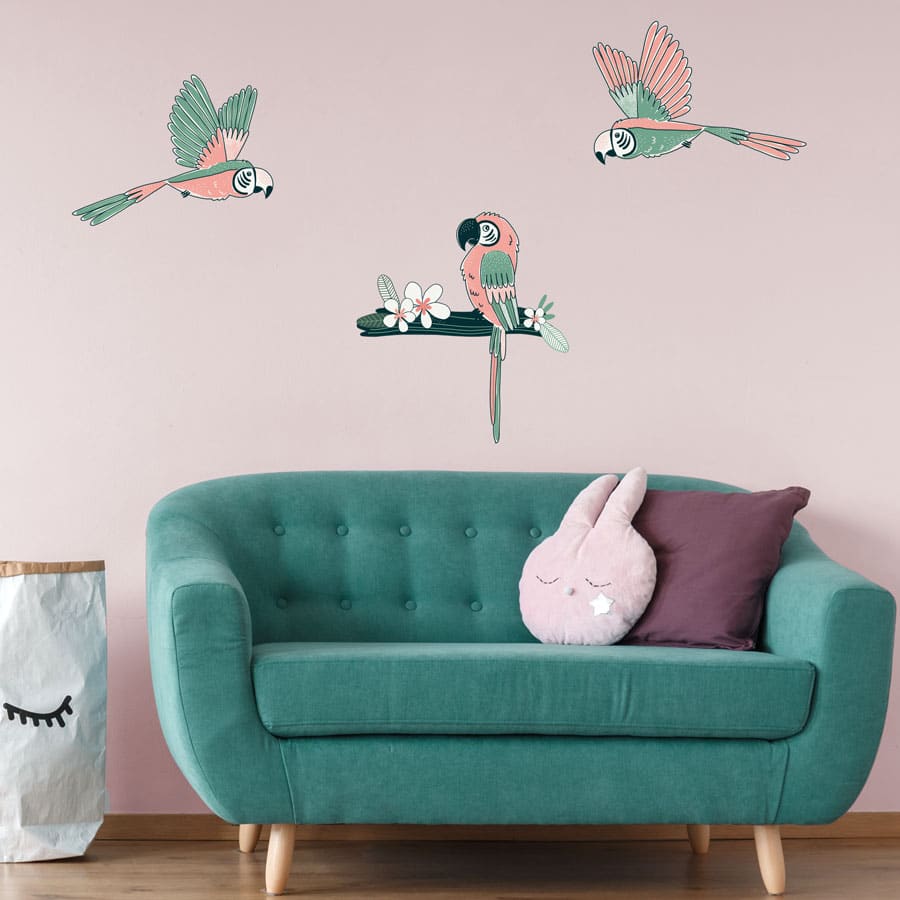 Tropical parrot wall sticker pack, jungle wall stickers. Image shows three pink and mint green parrot stickers, two are flying and one is sat on a dark green branch with a white flower. The stickers have been placed on a pale pink wall above a green sofa.