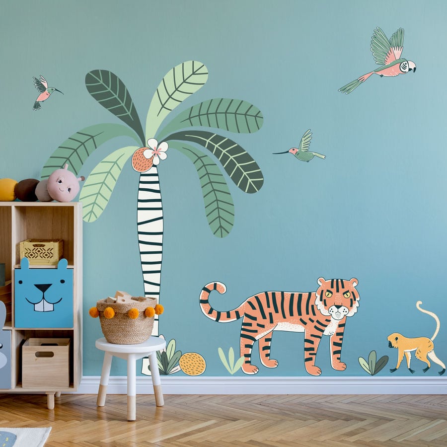 jungle wall sticker pack, jungle wall stickers. Image shows a large striped tree trunk with bright green leaves and coconuts, a tiger, monkey, a parrot and two smaller birds. The sticker has been placed on a light blue wall behind a small bookcase and stool.