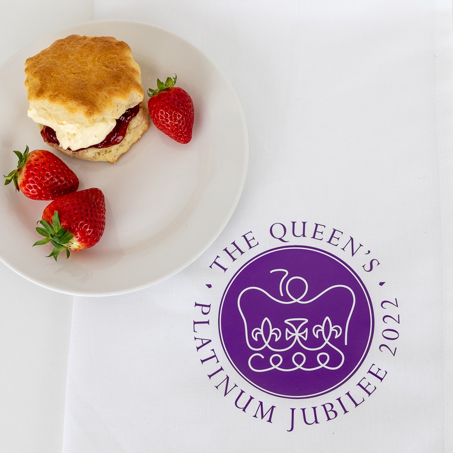 Official platinum jubilee logo tea towel shown flat with a plate of scones and strawberries