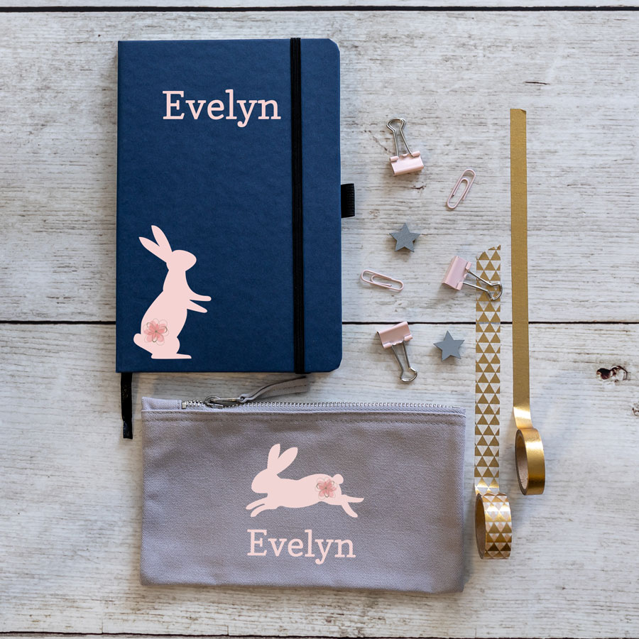 bunnies back to school mini bundle. Blue notebook with pink rabbit on bottom left with name in large pink text middle top. Grey pencil case with leaping pink rabbit in centre with name text in pink below.
