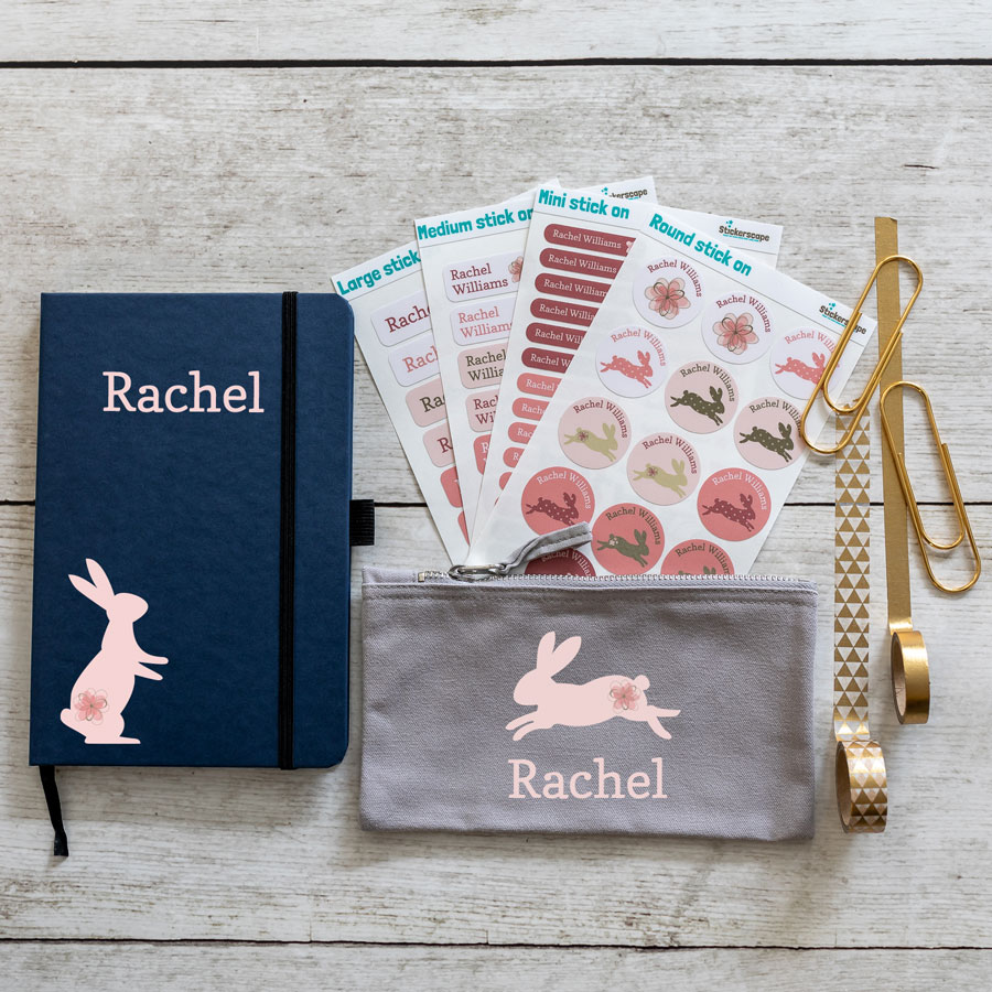 bunnies back to school mini bundle. Blue notebook with pink rabbit on bottom left with name in large pink text middle top. Grey pencil case with leaping pink rabbit in centre with name text in pink below. 4 sheets of stick on name labels.