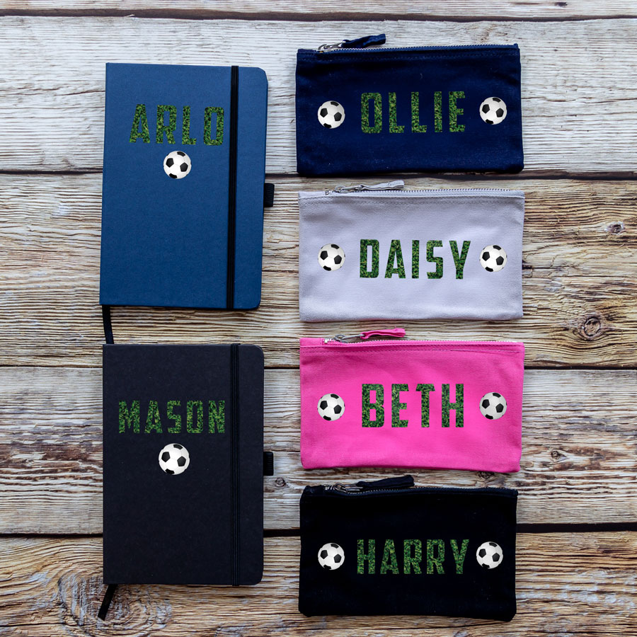Colour options for back to school bundle. A blue notebook, black notebook with the name in grass texture above a single football. A navy pencil case, grey pencil case, pink pencil case and black pencil cases with name in a grass print between two footballs.