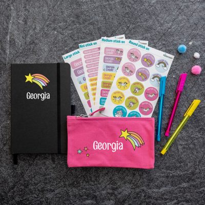rainbow shooting star back to school mega bundle. black notebook with rainbow shooting star image above name text in white. pink pencil case with white name text in centre, shooting star to top right and three stars bottom left. 4 sheets of stick on name labels.