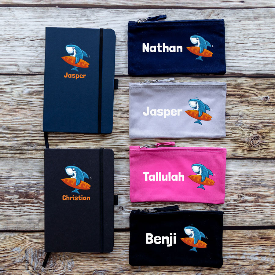 Little Shark back to school bundle colour options. Blue and black notebook, navy, grey, pink and black pencil case. Image of blue shark holding orange surf board with name text underneath.