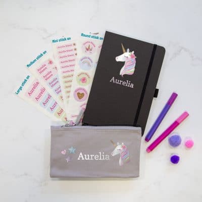 Unicorn back to school mega bundle featuring four pages of stick on name labels, a black notebook with white name text below a pastel pink unicorn head and and grey pencil case with name in white and centre with unicorn to the right and a small heart and two small stars to the left.
