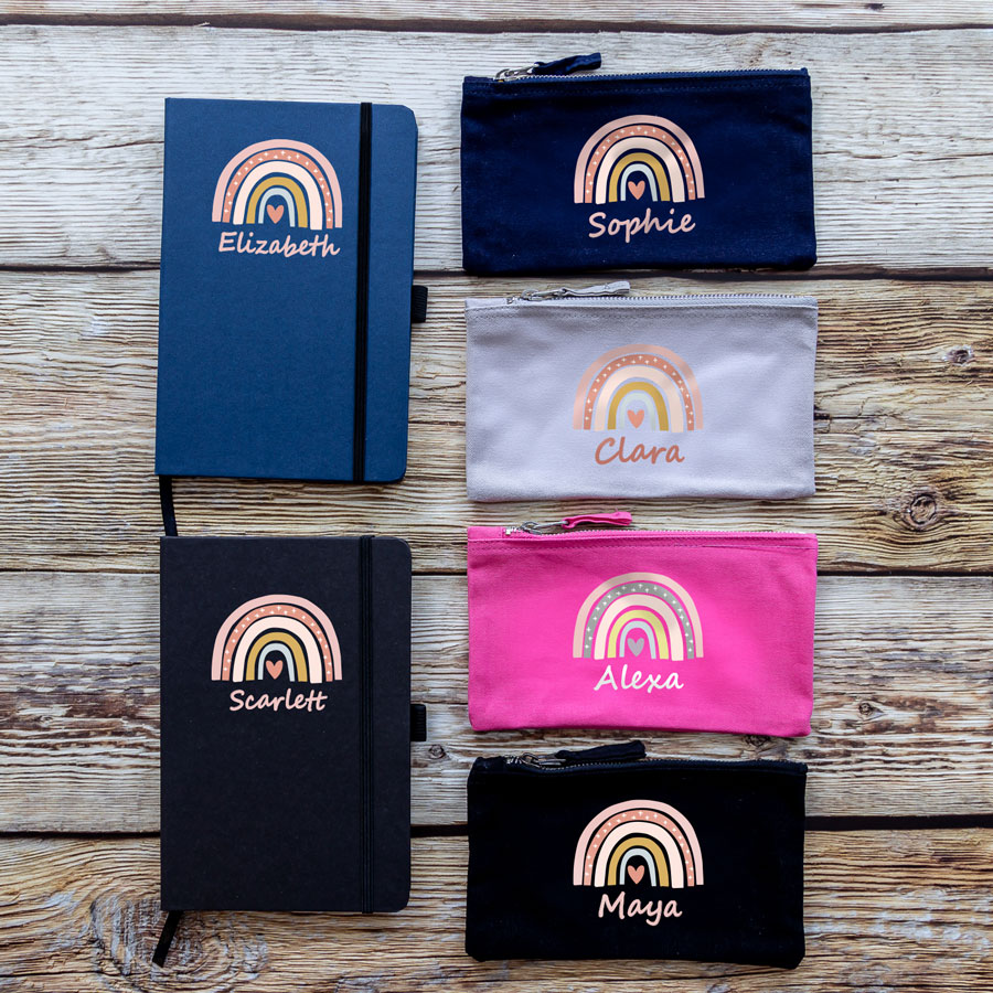 Rainbow heart back to school mini bundle colour options. Blue notebook, black notebook, navy pencil case, grey pencil case, pink pencil case, black pencil case. All with a pastel pink toned rainbow with heart in centre and pink coloured text below.
