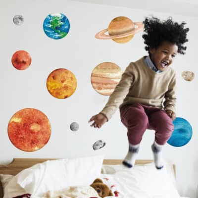 A joyful child jumping on a bed in front of colourful watercolour-style solar system wall stickers featuring planets, the sun, moon, and asteroids on a white wall.