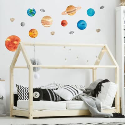 Watercolour-style solar system wall stickers featuring planets, the sun, moon, asteroids, and comets on a white wall in a modern kids' bedroom with a wooden house-frame bed.
