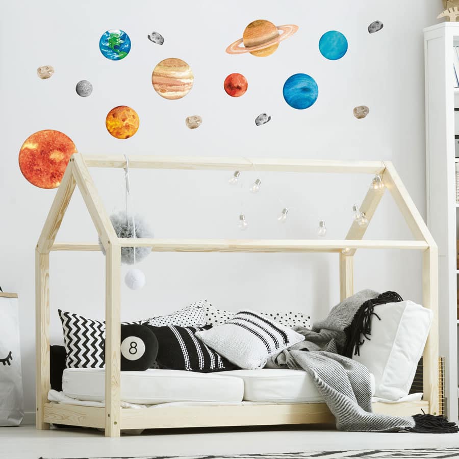 Watercolour-style solar system wall stickers featuring planets, the sun, moon, asteroids, and comets on a white wall in a modern kids' bedroom with a wooden house-frame bed.