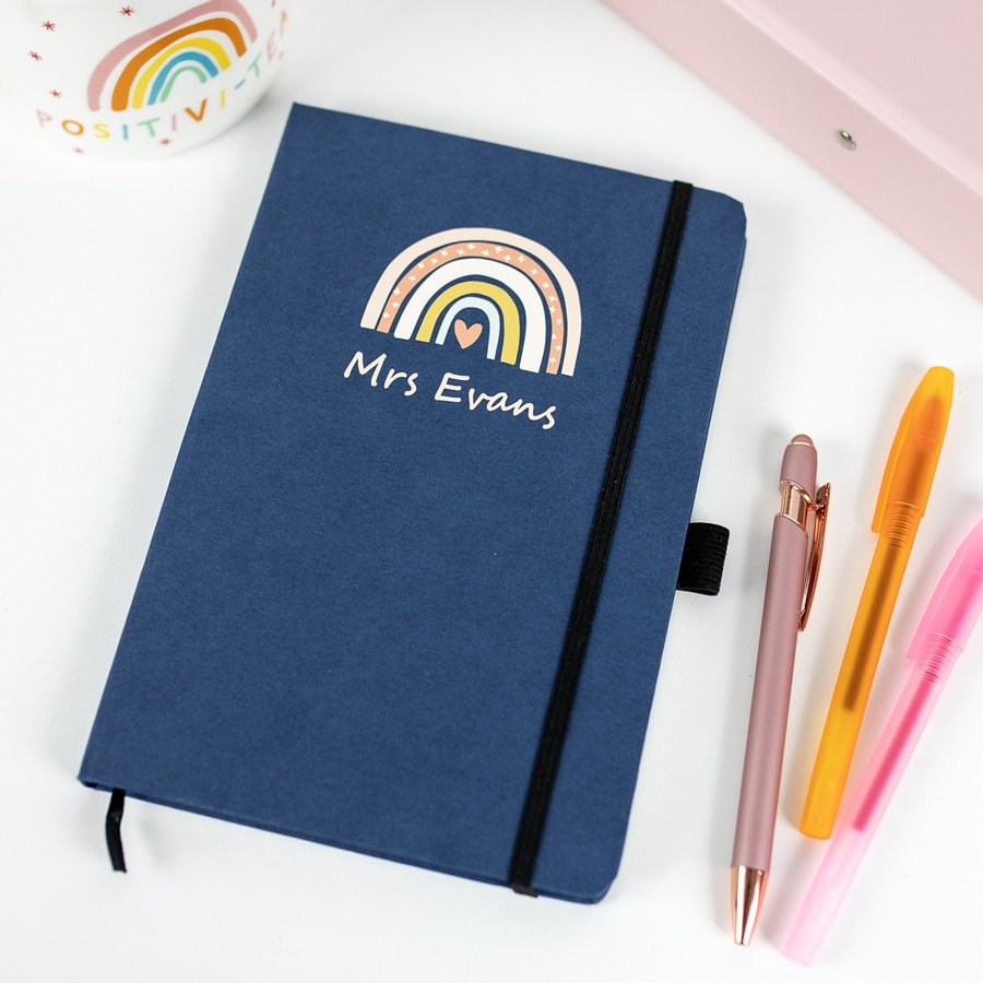 personalised rainbow notebook in blue with teacher accessories around the notebook on a white table