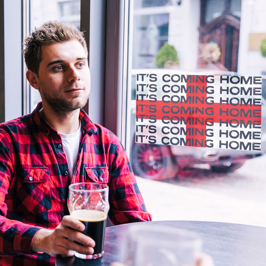 It's coming home window sticker option 2 shown on a window behind a man drinking a pint of beer at a table