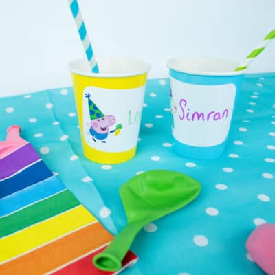 Peppa pig birthday label pack peppa & friends shown on two paper cups on top of a colouful table cover