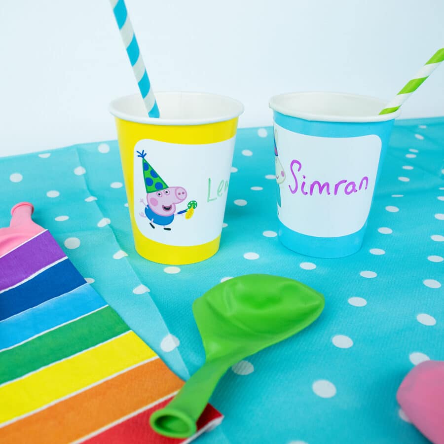 Peppa pig birthday label pack peppa & friends shown on two paper cups on top of a colouful table cover