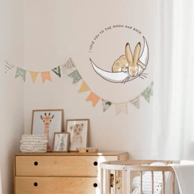 To the moon and back wall sticker shown on a light cream wall above pastel pink and green bunting and a light wooden dresser and cot
