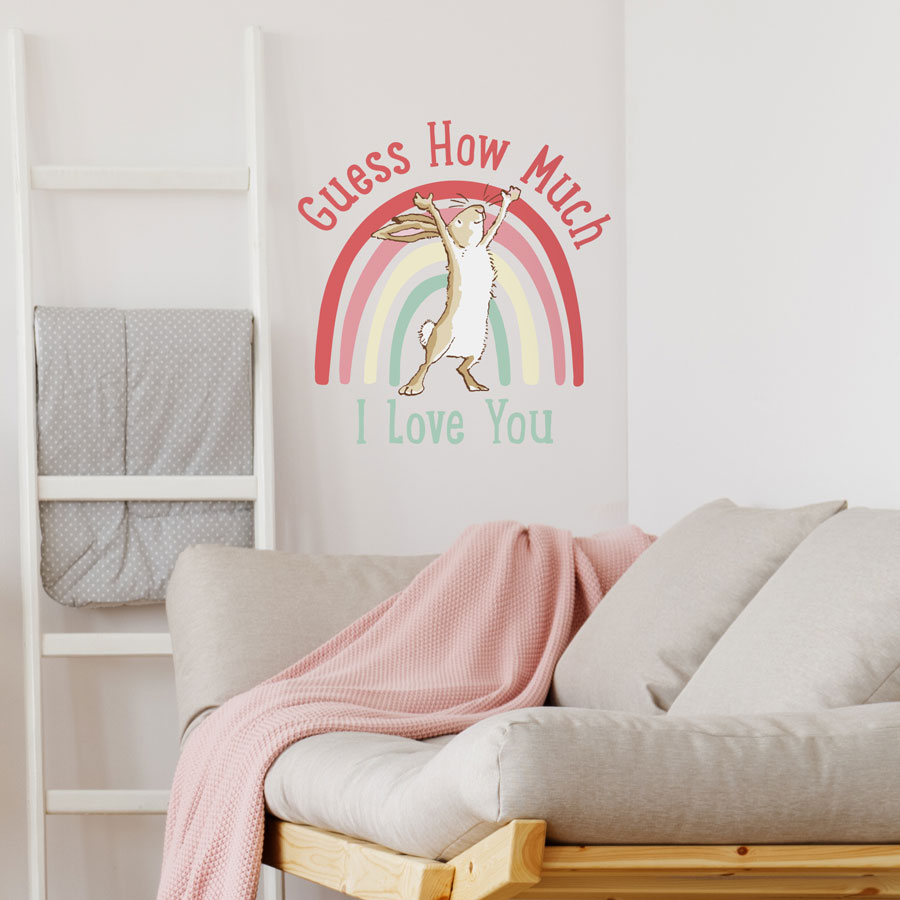 Rainbow hare wall sticker with text shown on a white wall behind a grey sofa with a pink blanket
