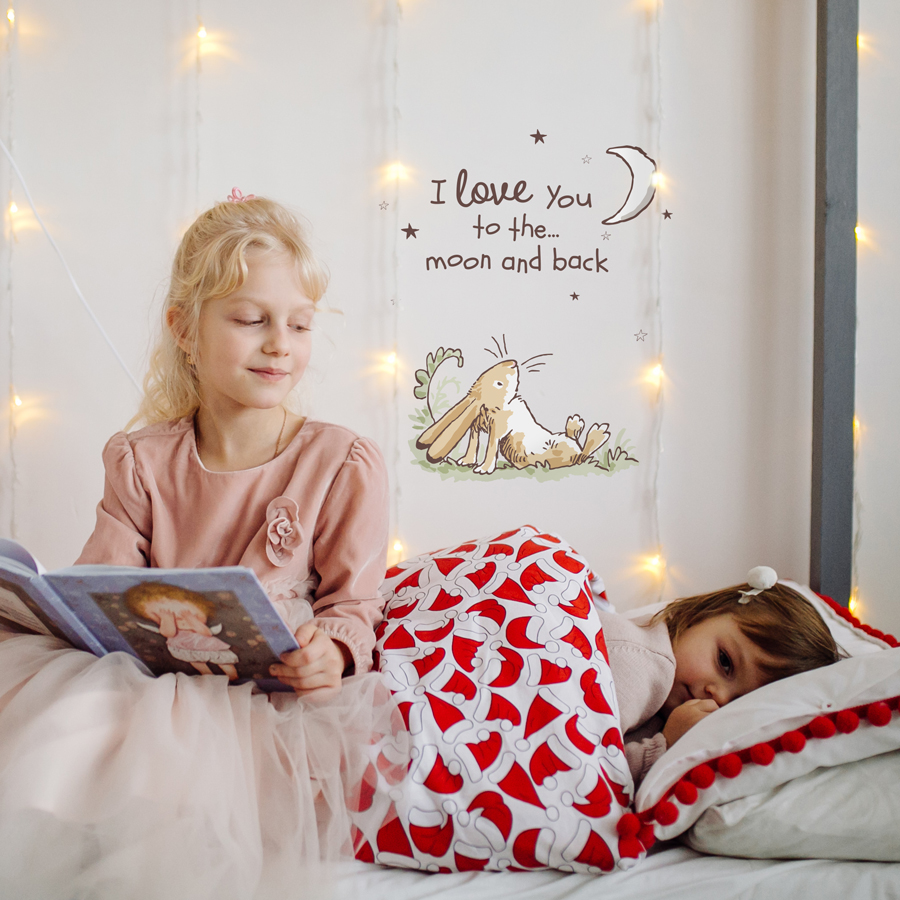 Moon & hare wall sticker regular shown on a white wall behind hanging fairy lights and two children reading a picture book