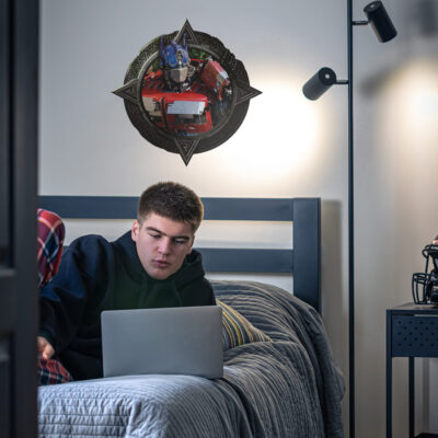 Optimus Prime wall sticker shown on a white wall above a bed with boy on laptop.