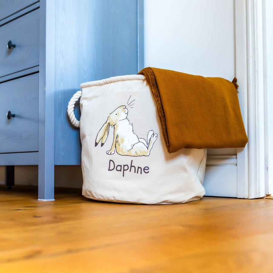 Canvas nursery storage basket with Guess How Much I Love You book inside and a mustard blanket draped over the side – ideal for baby essentials.