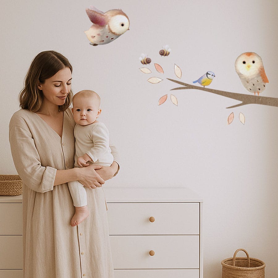 Nursery scene with woodland branch wall stickers on the wall, showing owls and bees, with mum holding baby in front of neutral dresser.