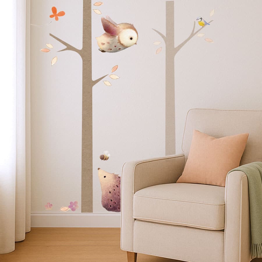 Neutral nursery corner with woodland tree wall stickers showing owl, bird, hedgehog and bees next to an armchair.